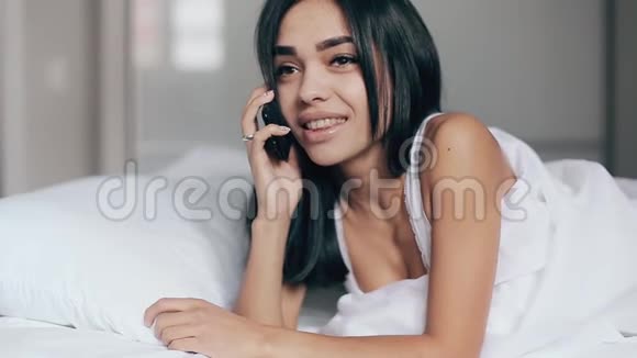 美丽的年轻黑发女人微笑着躺着说着智能手机在卧室里放松视频的预览图