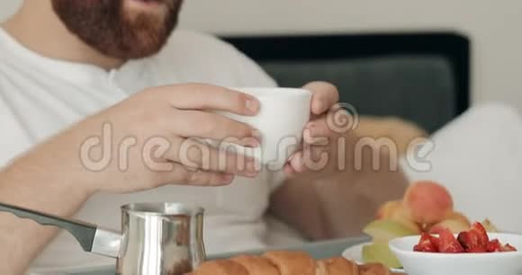 在床上吃早餐的时候英俊的留着胡须的男人一边喝着早咖啡三十岁的家伙拿着杯子视频的预览图