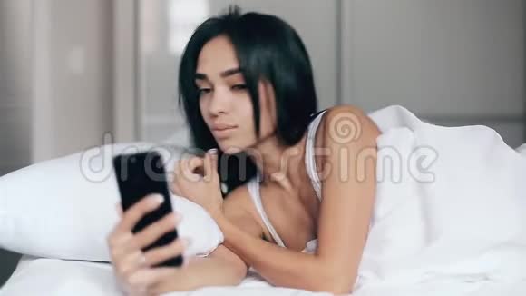 一个漂亮的女孩在床上用智能手机自拍视频的预览图