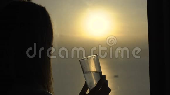 一位年轻女子在一间公寓里可以看到海岸和日出的全景她喝水视频的预览图