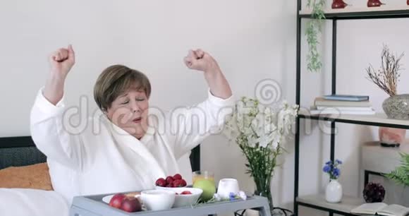 在罗伯的老快乐女士在早上吃早餐时伸展身体看着镜头成熟的美女坐着视频的预览图
