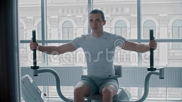 现代健身房一个穿白色t恤的年轻人在训练器械上做手臂运动视频的预览图