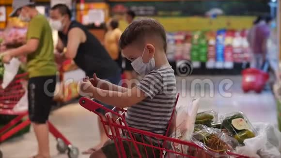 一家人在超市购物一个戴着防病毒面具的孩子坐在购物车里看电视视频的预览图