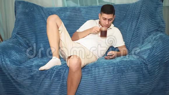 年轻人躺在沙发上看着电话喝着茶浏览社交网络查看电子邮件视频的预览图