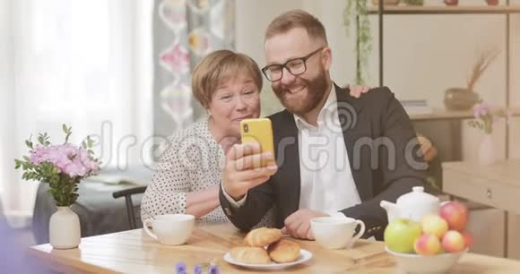 开朗的成年儿子和成熟的母亲坐在家里用智能手机打视频电话老年妇女和青年视频的预览图