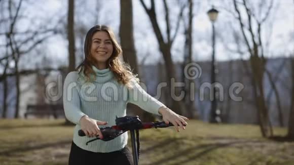 穿着绿松石毛衣的开朗的年轻女人拿着电动滑板车在春天公园笑慢动作视频的预览图