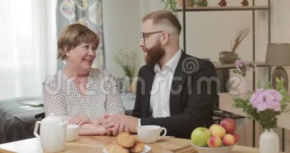 快乐成熟的母亲和成年的儿子在一起坐在厨房里聊天和放松年轻的商人来拜访她视频的预览图