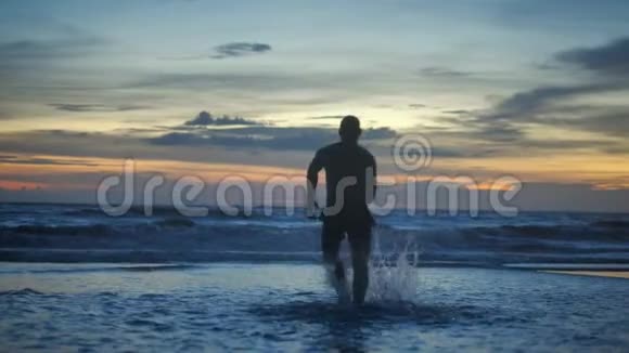 一个肌肉发达的男人的剪影在日落时赤裸的躯干在海滩上行走视频的预览图