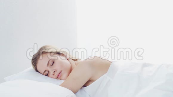 年轻的女人躺在床上拿着智能手机美丽的金发觉醒女孩早上在卧室里白天视频的预览图