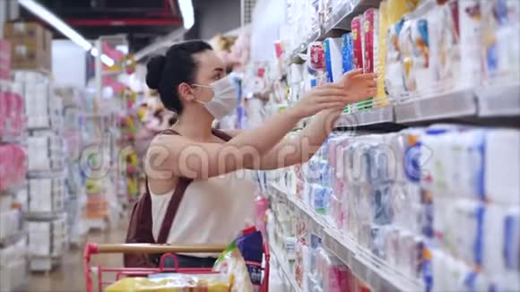 一位戴着冠状病毒流行面具的年轻女子站在超市里人们在那里惊慌失措地购买视频的预览图