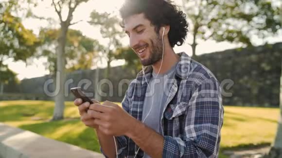 一位笑容可掬的年轻人用手机在公园里发短信他的耳朵里插着白色耳机视频的预览图