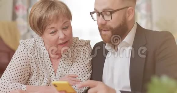 英俊的商人用现代智能手机教他成熟的祖母漂亮的老妇人和年轻人视频的预览图