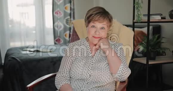 好看的老太太坐在椅子上在家里支撑着头的肖像美丽的退休妇女微笑视频的预览图