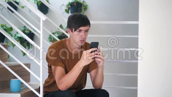悲伤的年轻人坐在家里的楼梯上用智能手机视频的预览图