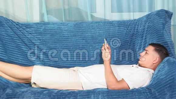 穿着白色t恤的年轻人正拿着电话躺在沙发上视频的预览图