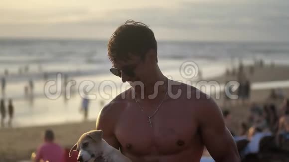 日落时分戴着太阳镜带着一只可爱的小狗在海滩上微笑的肌肉男的肖像视频的预览图