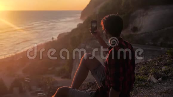 年轻人的剪影在他的智能手机上制作美丽日落的照片或视频旅游博主制作社交照片视频的预览图