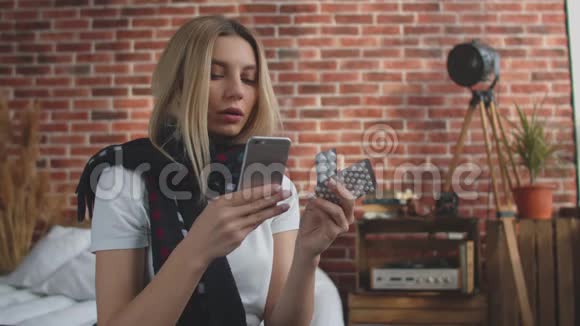 一个脖子上戴着围巾的生病的女孩坐在床上读着使用药片的说明女孩坐在视频的预览图