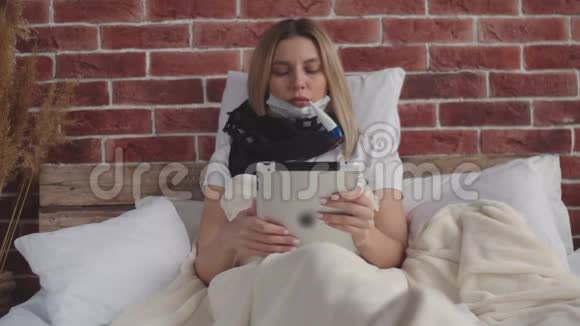 一个戴着围巾的生病女孩躺在床上手里拿着一块数字平板电脑拿出一个温度计视频的预览图