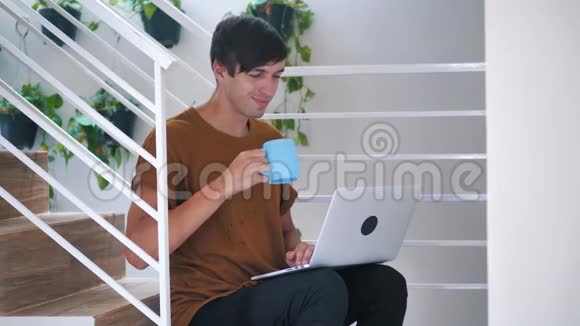 年轻人在家里坐在楼梯上用笔记本电脑和喝咖啡视频的预览图