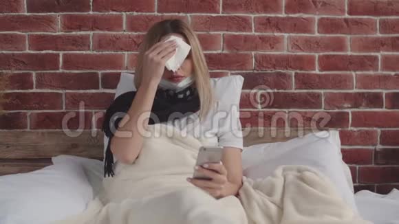 一个戴着围巾的生病女孩躺在床上手里拿着手机擦着汗视频的预览图