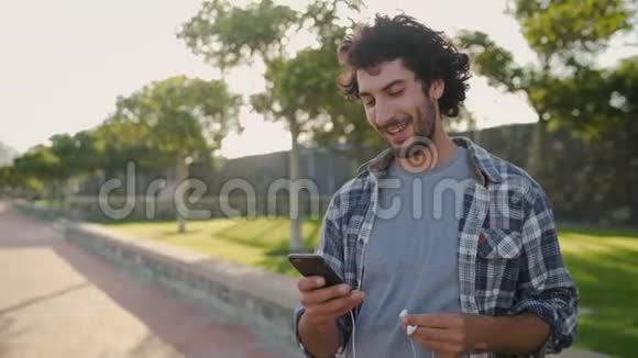 一位年轻人在公园里用手机在耳朵里插入耳机并在网上发短信时的微笑画像视频的预览图