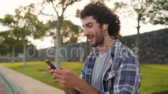 一位英俊快乐的年轻人戴着耳机在公园里享受智能手机上发短信的肖像快乐视频的预览图
