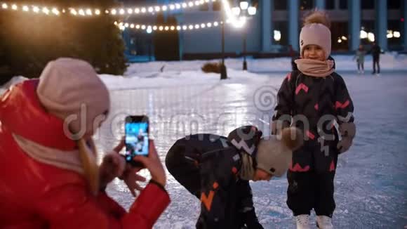 两个小孩在冰场上他们的母亲用电话摄影机拍摄他们视频的预览图