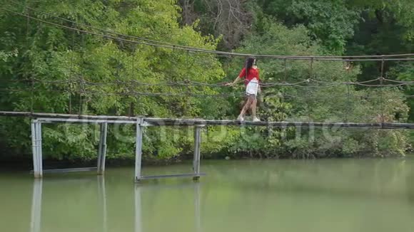 穿着红色衬衫和白色裙子的性感年轻黑发女人在画报公园里的河上悬桥散步视频的预览图
