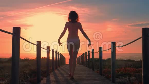 日落时分一位年轻女子在沙滩上的木脚桥上剪影视频的预览图