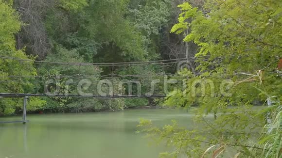 穿着红色衬衫和白色裙子的性感年轻黑发女人在画报公园里的河上悬桥散步视频的预览图