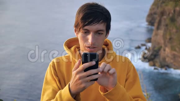 年轻的徒步旅行者站在山上在悬崖和海浪的背景下滚动手机视频的预览图