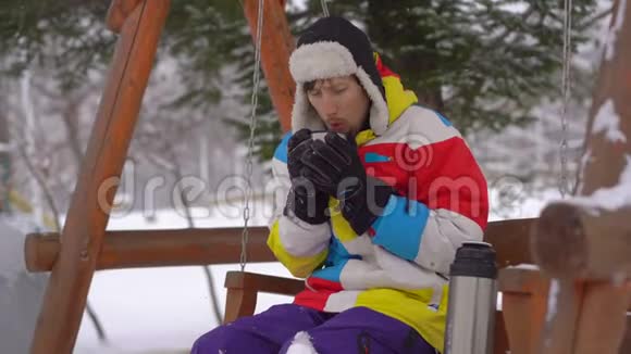 降雪期间的年轻人坐在长凳上喝热饮料他觉得很冷寒冷天气概念视频的预览图