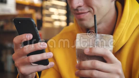 现代咖啡馆的智能手机触摸屏为穿黄色连帽衫的男性特写喝奶昔或冰沙视频的预览图