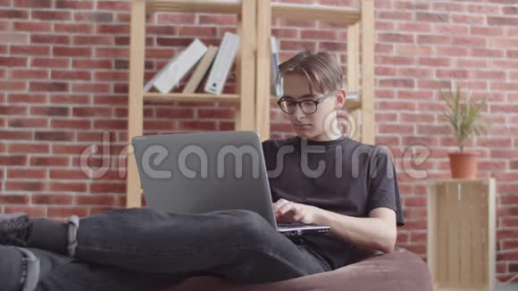 那家伙坐在一张包椅上在笔记本电脑上打字对着相机微笑一个穿鼻子的年轻臀部视频的预览图