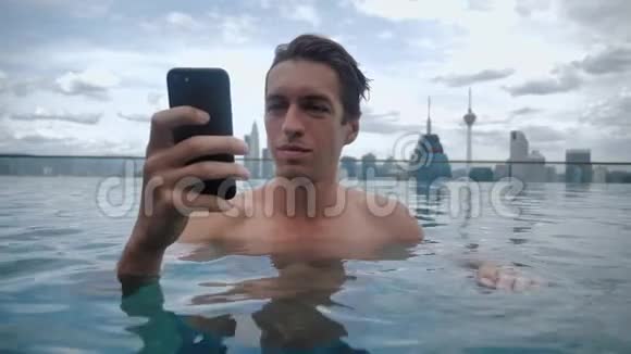 年轻人用智能手机在城市背景下摩天大楼屋顶上的游泳池里放松视频的预览图