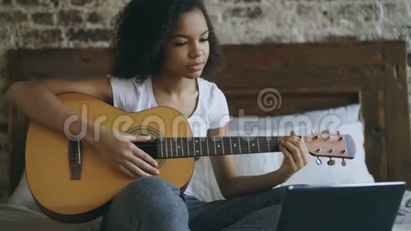 年轻的卷发混血女孩集中学习弹吉他使用笔记本电脑坐在家里的床上视频的预览图