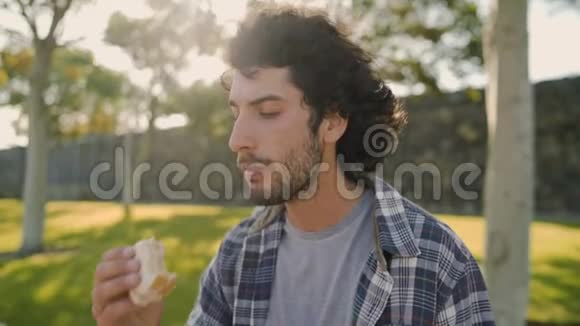 阳光明媚的一天一个英俊的微笑的年轻人坐在公园里吃三明治在公园里吃午饭视频的预览图