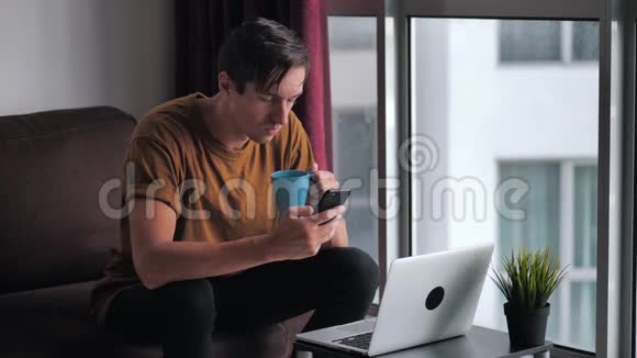忙碌的年轻人一边坐在沙发上靠窗一边用智能手机和笔记本电脑喝咖啡视频的预览图