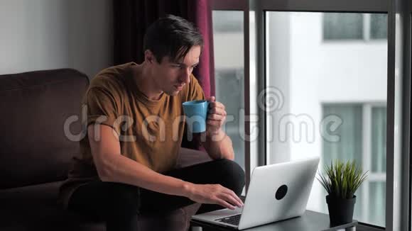 英俊的年轻人一边喝咖啡一边用笔记本电脑坐在家里的沙发上窗前视频的预览图