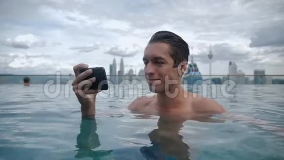 年轻人在游泳池里放松在城市背景下摩天大楼屋顶上的智能手机上观看视频视频的预览图
