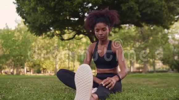 一位非洲裔美国青年妇女坐在绿草上在公园跑步前伸展双腿的肖像视频的预览图