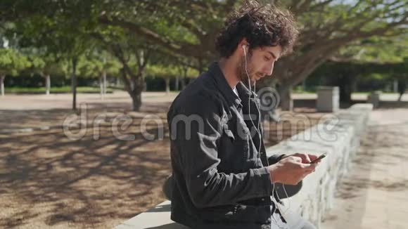 一位年轻快乐的年轻臀部滑板运动员的画像他的耳朵里戴着耳机在手机上发短信视频的预览图
