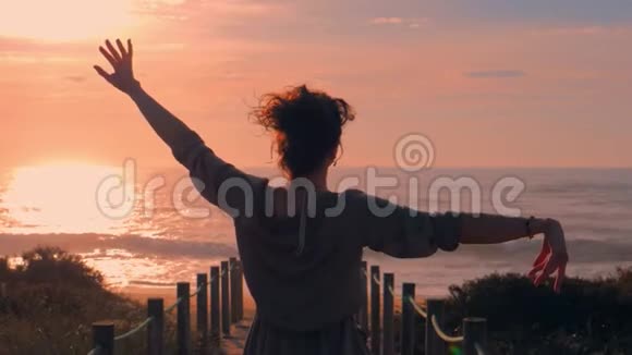 日落时分一位女士双臂走上沙滩上的木脚桥视频的预览图