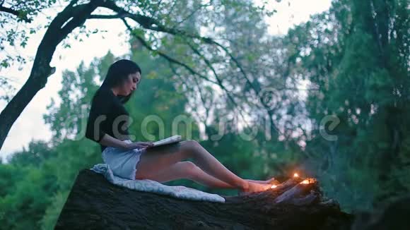 迷人的性感年轻黑发女人坐在倒下的树干上看书靠近她的脚烧蜡烛视频的预览图