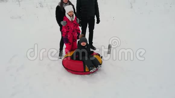 一家人站在森林附近的雪田中间那个小男孩坐在充气雪橇上视频的预览图