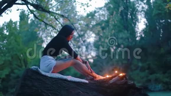 迷人的性感年轻黑发女人坐在倒下的树干上看着她脚附近燃烧的蜡烛视频的预览图