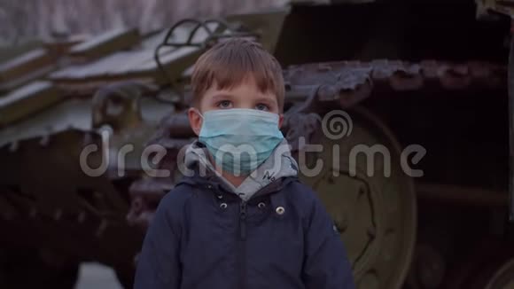 戴医用防护面罩的男孩在COVID19大流行爆发时站在军用机器站立并观察视频的预览图
