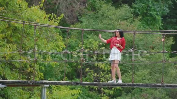 身着鲜红色衬衫和白色裙子的年轻黑发女子站在河边的吊桥上自拍视频的预览图