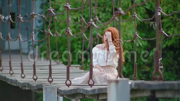迷人的可爱的年轻女子一头火红的头发坐在河上的吊桥上想着什么视频的预览图